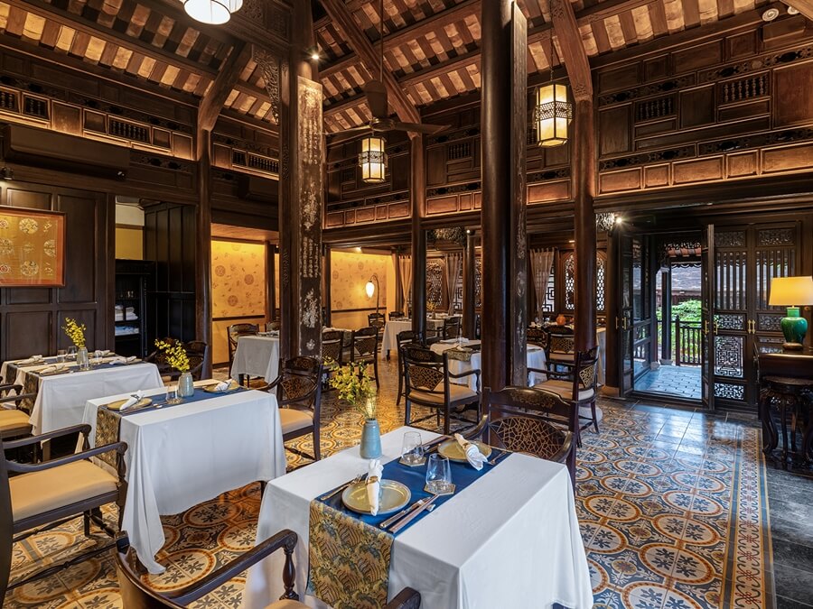 Wide shot of the main dining hall, showcasing towering dark wood pillars, high vaulted ceilings, warm hanging lanterns, and colourful vintage patterned floor tiles