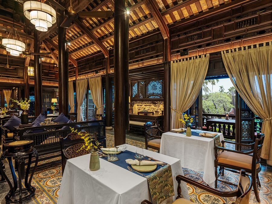 Interior view of the restaurant showing multiple dining tables, heavy wooden structure, soft cream curtains framing a tropical garden view, and ornate wooden carvings in the background.