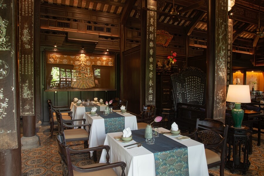Close-up of a dining corner in a traditional Hue garden house restaurant, featuring dark carved wood, mother-of-pearl inlay, set tables with white linen and blue runners, and a classic tiled floor