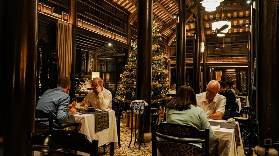 Diners seated at white-clothed tables within the wood-paneled hall, centered around a subtly decorated Christmas tree, emphasizing the warm, intimate lighting