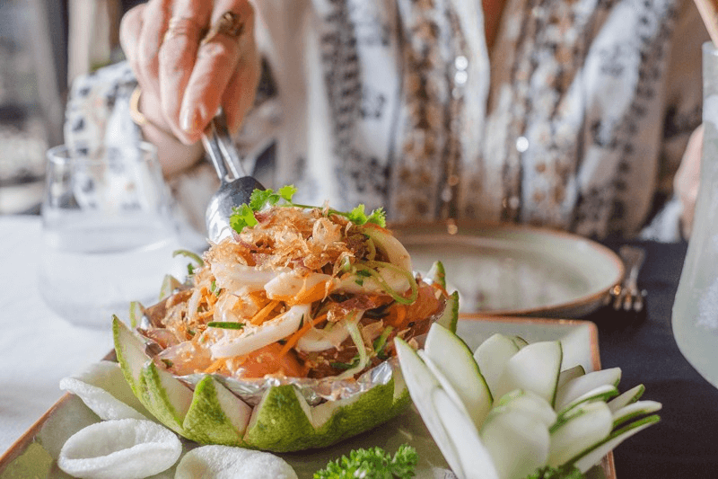 Close-up of a beautifully carved pomelo shell holding a fresh Vietnamese salad, garnished with fried shallots and herbs, with a hand reaching in with a fork