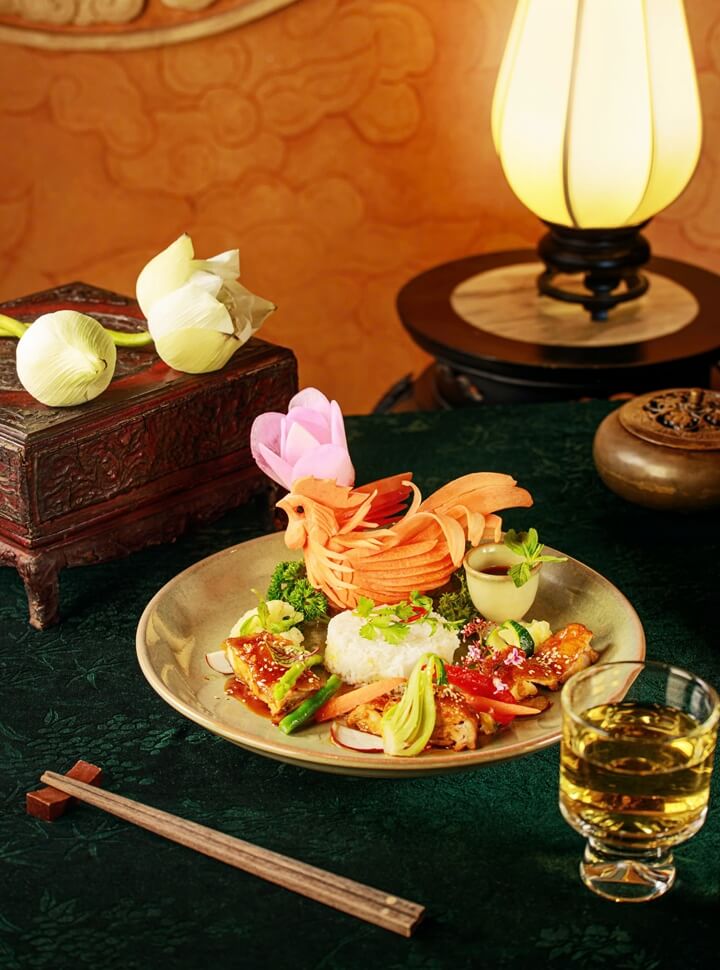 Meticulously plated main course with a portion of rice, glazed chicken, and vegetables, highlighted by a large, elaborate phoenix or rooster carving made from a vegetable, on a dark green table setting