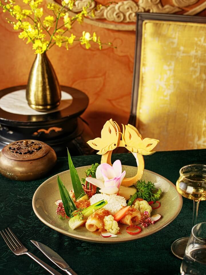 Gourmet Vietnamese dish featuring rice and squid, garnished with vegetables and a delicate carved butterfly decoration, set on a dark green tablecloth next to a brass vase and incense burner