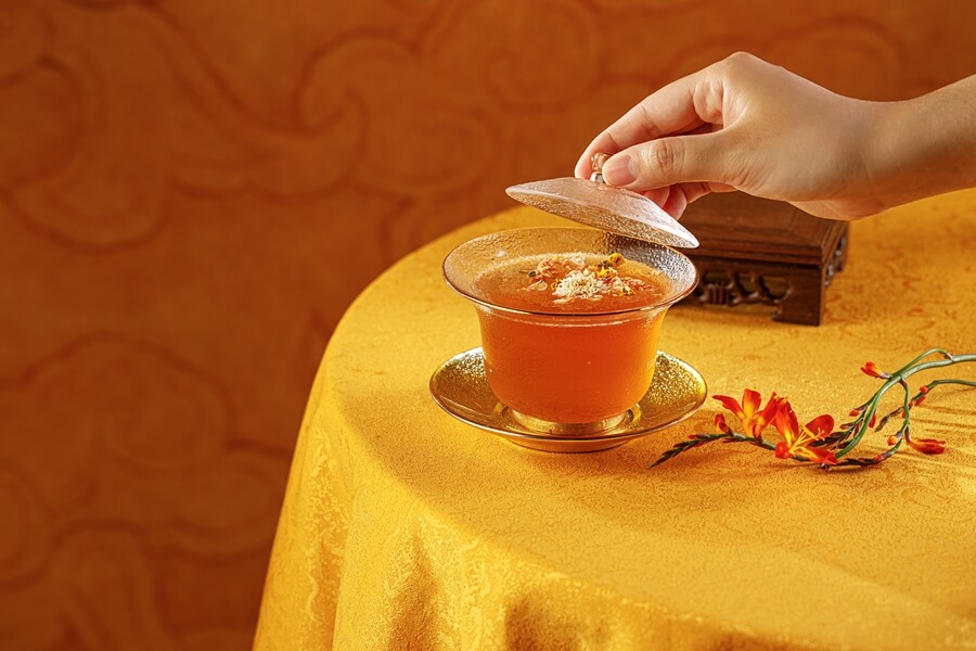 A gentle hand lifts the lid from a clear glass cup of warm, amber-coloured Chrysanthemum Tea, signifying a delicate serving ritual on a golden, ornate tablecloth