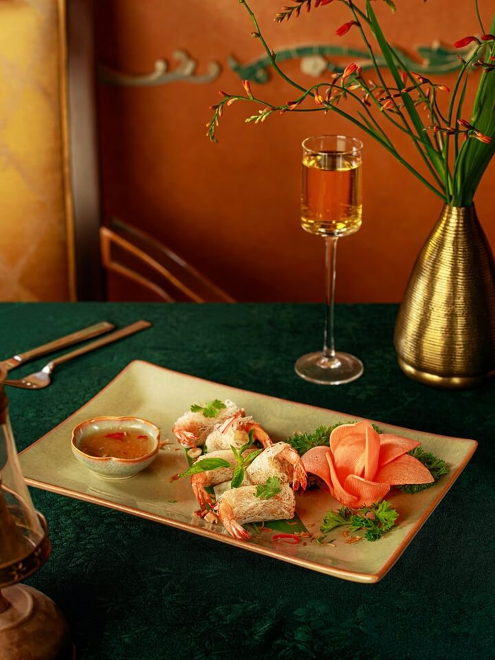 An appetizer of crispy prawn rolls served on a rectangular platter with a side of dipping sauce, meticulously garnished with a sculpted flower shape, next to a tall glass of amber-coloured beverage